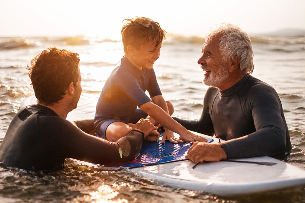 Tre generasjoner deler et rolig øyeblikk på et surfebrett i sjøen ved solnedgang.